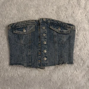 Jeans tubtopp med knappar - Snygg jeans tubtopp med knappar framtill och två dekorativa fickor. Perfekt för en trendig look. Den har en rå kant nedtill för en avslappnad stil.