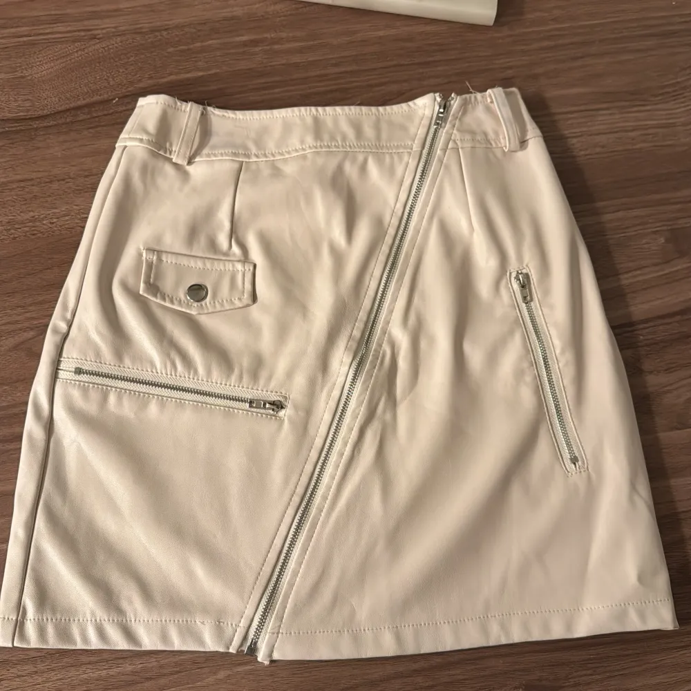 Snygg beige kortkjol med en unik sned dragkedja framtill och praktiska fickor med dragkedjor. Kjolen har bälteshällor och en stilren design som passar perfekt för en trendig look.. Hameet.