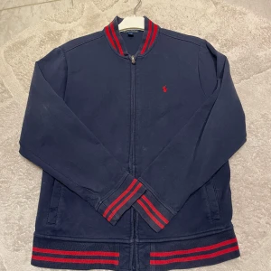 Blå jacka från Polo Ralph Lauren - Säljer en snygg blå jacka från Polo Ralph Lauren med röda detaljer. Jackan har dragkedja och långa ärmar med ribbade muddar. Perfekt för en stilren look. 18-20