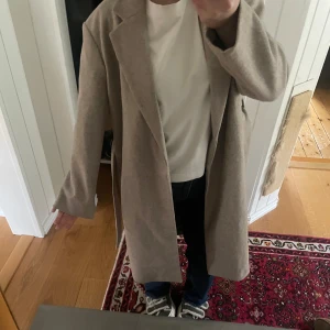 Vågig beige lång kappa   - Säljer en stilren beige brun kappa från ONLY i storlek XS. Kappan har en klassisk design med knytbälte i midjan och långa ärmar. Perfekt för att hålla stilen under kyligare dagar. Helt ny