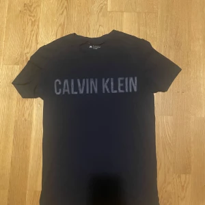 Mörk blå t-shirt från Calvin Klein - Snygg mörkblå t-shirt från Calvin Klein med deras ikoniska logga tryckt på bröstet. Perfekt för en stilren och avslappnad look. T-shirten är kortärmad och gjord i mjukt material för extra komfort.