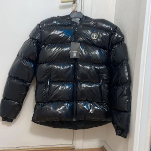 Svart pufferjacka från Trapstar - Säljer en svart pufferjacka från Trapstar med glansig finish och broderad logga på ryggen. Jackan har dragkedja framtill och långa ärmar. Perfekt för kyliga dagar när du vill hålla stilen. 🖤