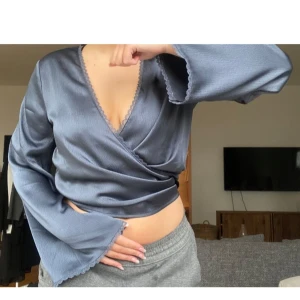 Blå omlottopp med knytband - Snygg blå omlottopp med långa ärmar och knytband i midjan. Toppen har en v-ringad design och är tillverkad i ett glansigt material som ger en elegant look. Perfekt för att skapa en stilren outfit.