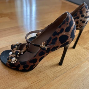 Leopardmönstrade klackskor från YSL - Snygga leopardmönstrade klackskor från YSL med dekorativa pärlor och blommor framtill. Skorna har en elegant rem över foten och en hög klack. Perfekta för att ge en djärv touch till din outfit.