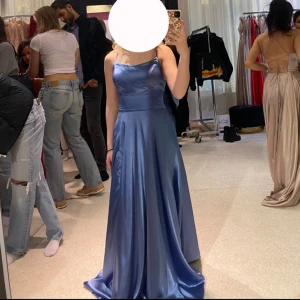 Balklänning för kort person - Inköpspris: 3300. Elegant blå långklänning i satin med smala axelband och v-ringad design. Klänningen har en öppen rygg med snygg snörning som ger en unik look. Perfekt till balen! Klänningen är använd 1 gång och visar små tecken på användning som knappt synligt. Klänningen är uppsydd och anpassad för en person på ca 156 cm lång.