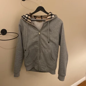 Grå hoodie från Burberry - Säljer en stilren grå hoodie från Burberry med klassiskt rutigt mönster i huvan. Tröjan har dragkedja framtill och är tillverkad i mjuk bomull. Perfekt för en avslappnad look.