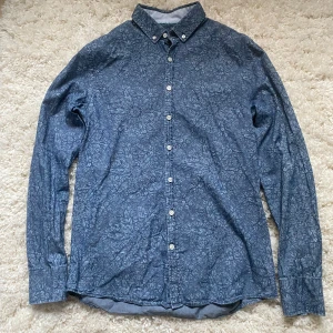 Blå blommig skjorta från Savvy Citizen - Snygg blå skjorta från Savvy Citizen med ett diskret blommigt mönster. Skjortan har långa ärmar och knappar framtill. Perfekt för en stilren look.