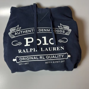 Mörkblå hoodie från Ralph Lauren - Säljer en snygg mörkblå hoodie från Ralph Lauren med tryck framtill. Den har en klassisk design med dragsko i huvan och ribbade muddar vid ärmslut och nederkant. Perfekt för en avslappnad stil.