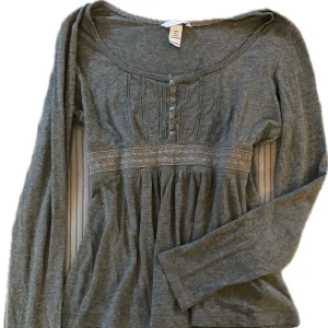 Grå långärmad topp med spetsdetaljer - Säljer en stilren grå långärmad topp från H&M. Toppen har fina spetsdetaljer under bysten och knappar framtill. Lätt urringad och super skönt bomull & viscos tyg. 
