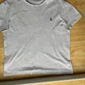 Grå t-shirt från Ralph Lauren - Snygg grå t-shirt från Ralph Lauren med klassisk broderad logga på bröstet. Perfekt för en stilren och avslappnad look. Tillverkad i mjukt bomullsmaterial för extra komfort.