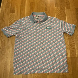 Randig pikétröja från Lacoste - Säljer en snygg randig pikétröja från Lacoste i ljusblått, rosa och vitt. Tröjan har korta ärmar och en klassisk krage med knappar. 