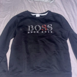Svart tröja från Hugo Boss - Säljer en stilren svart tröja från Hugo Boss med logotypen tryckt på framsidan. Tröjan har en klassisk rund hals och långa ärmar. Perfekt för en avslappnad och trendig look.
