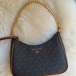 Brun axelväska från Michael Kors - Säljer en superfin brun axelväska från Michael Kors!  ✔️ Klassisk design med MK-mönster ✔️ Guldiga detaljer och kedja för en lyxig touch ✔️ Perfekt storlek – rymmer det viktigaste utan att vara klumpig ✔️ I mycket fint skick!  Passar både till vardag och fest, och är en tidlös accessoar som lyfter vilken outfit som helst. Skicka ett meddelande vid intresse! ✨ det är små fläckar på insidan av väskan, som en bild visar! 