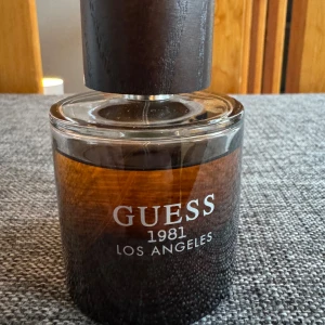 Guess 1981 Los Angeles Parfym - Säljer en elegant parfym från Guess, 1981 Los Angeles. Flaskan är rund med en stilren design i brunt glas och har en matchande brun kork. Perfekt för den som vill ha en sofistikerad doft med en touch av Los Angeles glamour. Mängden enligt bilden cirka 90% av 100ml.