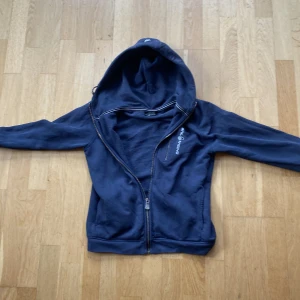 Blå Hoodie från Sail Racing - Säljer en blå fleecejacka från Sail Racing med dragkedja och huva. Jackan har en bröstficka med dragkedja och märkeslogga på bröstet. Perfekt för kyliga dagar!