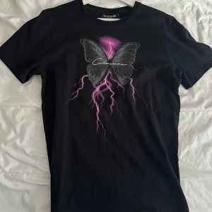 Svart t-shirt med fjärilsmotiv från Closure - Cool svart t-shirt från Closure med ett stort fjärilsmotiv i mitten. Fjärilen är omgiven av lila blixtar som ger en häftig effekt. Perfekt för dig som vill sticka ut med en unik design.