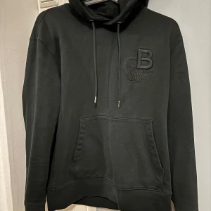 Svart hoodie från Boss - Säljer en stilren svart hoodie från Boss med en broderad logga på bröstet. Den har en klassisk känguruficka och justerbar huva med snören. Perfekt för en avslappnad look. Den är även sällsynt och går inte att hitta på marknaden längre