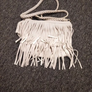 Vit axelväska med fransar från H&M - Snygg vit axelväska från H&M med coola fransdetaljer. Väskan har en flätad axelrem och stängs med dragkedja. Perfekt för att ge din outfit en bohemisk touch.