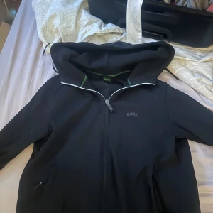 Svart jacka med huva från BOSS - Säljer en stilren svart jacka från BOSS med huva och dragkedja framtill. Köpt för 2000kr säljs för 1000kr