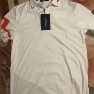 Vit pikétröja från J.Lindeberg - Snygg vit pikétröja från J.Lindeberg med röda detaljer på kragen och ärmen. Perfekt för en stilren look. Tröjan har en klassisk krage och korta ärmar. Ny pris ca800kr 