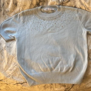 Ljusblå tröja med pärlor från Zara - Säljer en ljusblå tröja från Zara med korta ärmar och dekorativa pärlor vid halsringningen. Tröjan har en ribbad kant vid ärmslut och nederkant, vilket ger en snygg passform. Aldrig använts