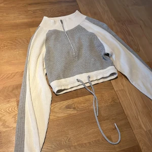 Grå och vit tröja från PrettyLittleThing - Säljer en snygg grå och vit tröja från PrettyLittleThing. Tröjan har en dragkedja framtill och en croppad design med snörning i nederkant. Perfekt för en stilren look.
