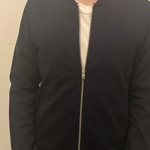 Mörkblå tröja från Lindbergh - Säljer en stilren mörkblå full-zip tröja från Lindbergh i slim fit. Tröjan har en dragkedja framtill och är tillverkad i ett mjukt material. Perfekt för en elegant look. (Kan Sicka dagen efter vid snabb affär)