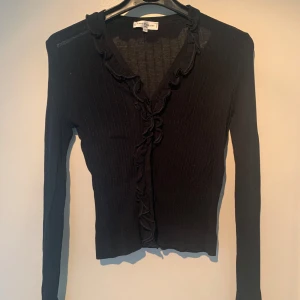 Svart ribbad kofta från Anne Fontaine - Elegant svart ribbad kofta från Anne Fontaine med volangdetaljer längs den v-ringade kanten. Perfekt för en stilren look. Långärmad design som passar bra till olika tillfällen. Går både att klä upp med kjol och ner med jeans! Franskt märke så liten i storleken (36/38)