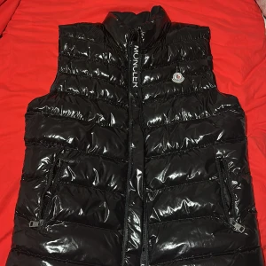 Svart dunväst från Moncler - Säljer en snygg svart dunväst från Moncler med glansig yta och dragkedja framtill. Västen har två praktiska fickor med dragkedjor och Moncler-logga på bröstet. Perfekt för kyligare dagar! Priset kan sänkas till 1000