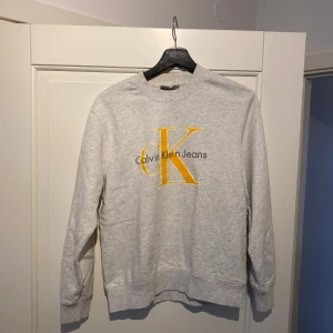 Grå sweatshirt från Calvin Klein Jeans - Säljer en stilren grå sweatshirt från Calvin Klein Jeans med ett stort gult CK-logotryck på bröstet. Tröjan har långa ärmar och en rund halsringning. Perfekt för en avslappnad look.