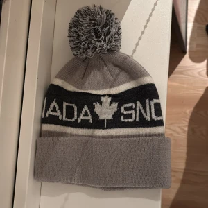 Grå mössa från Canada Snow - Snygg grå mössa från Canada Snow med text och lönnlövsmotiv. Den har en stor tofs på toppen och är tillverkad i en ull- och akrylblandning för extra värme. Perfekt för kyliga dagar! Nästintill aldrig använd. Inga skador eller märken. Säljs för jag redan har flera mössor. Köptes för 500 kr.