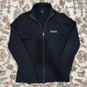 Svart tröja med dragkedja från Boss - Snygg svart tröja från Boss med dragkedja och broderad logga i guld på bröstet. Tröjan har en hög krage och långa ärmar, perfekt för en stilren look. Passar bra till både jeans och chinos.