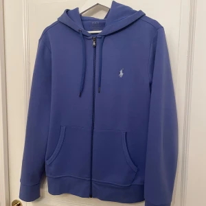 Baby Blå Ralph Lauren Zip Hoodie - Säljer en snygg blå hoodie från Ralph Lauren med dragkedja och huva. Den har en klassisk design med broderad logga på bröstet och praktiska fickor framtill. Perfekt för en avslappnad stil. Skick 9/10 lite solbränd där bak