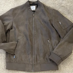 Brun bomberjacka från Jack & Jones - Säljer en snygg brun bomberjacka från Jack & Jones. Jackan har dragkedja framtill och på fickorna, samt en extra ficka på ärmen. Perfekt för en stilren look.