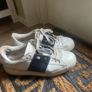 Valentino skor - Säljer ett par vita sneakers med svarta detaljer och snörning. Skorna har en cool design med nitar på hälen och en bred svart rem över sidan. Perfekta för en avslappnad och stilren look. Storlek 36,5 köpta för 3400 på plick