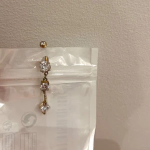 Guld Piercing med glittrande stenar - Säljer ett elegant guld piercing med tre glittrande stenar i olika storlekar.  Perfekt för att lysa upp vilken outfit som helst! (Köpt för ca 500), rostfritt stål och knappt använd. 