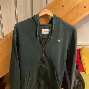 Säljer en snygg grön hoodie från Lyle & Scott i storlek M. Den är gjord av ekologiskt material och har en dragkedja framtill. Perfekt för en avslappnad stil med sin klassiska design och det ikoniska märket på bröstet. Passar både till vardags och chillkvällar. 🐥