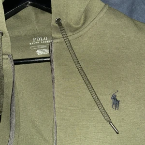 Olivgrön hoodie från Ralph Lauren - Säljer en snygg olivgrön hoodie från Ralph Lauren.Den har en dragkedja framtill och en klassisk känguruficka.Perfekt för höst och vinter med sin mjuka och bekväma passform. Den ikoniska Ralph LaurenLoggan är broderad på bröstet. Helt ny endast provad