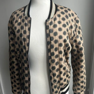 Beige prickig bomberjacka från Isabel Marant Étoile - Säljer en supercool beige bomberjacka från Isabel Marant Étoile med svarta prickar över hela. Jackan har en broderad text 'Africa' i rött på ryggen. Den är gjord av 100% bomull med en polyesterfyllning och ribbade kanter. Perfekt för att sticka ut i mängden!