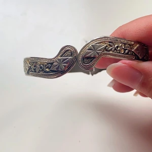 Vintage silverfärgat armband med detaljer - Säljer ett supercoolt vintage armband i silverfärg. Det har snygga ingraverade mönster och en unik design som verkligen sticker ut. Perfekt för att ge din outfit en retro touch! Armbandet är i bra skick och passar de flesta handleder.