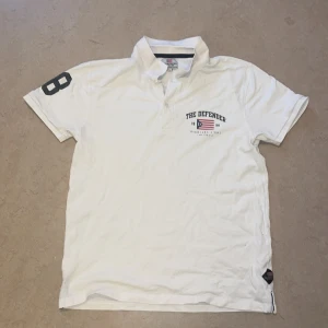 Vit pikétröja med tryck - Säljer en snygg vit pikétröja med texten 'The Defender' och ett coolt tryck på bröstet. Tröjan har korta ärmar och en klassisk krage. Perfekt för en avslappnad stil!