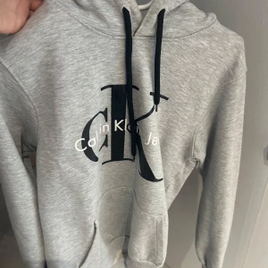 Grå hoodie från Calvin Klein - Säljer en snygg och bekväm grå hoodie från Calvin Klein. Den har en stor CK-logga på framsidan och en praktisk känguruficka. Perfekt för både vardag och chill. Huvan är fodrad med ett mörkare tyg för extra stilpoäng. Passar perfekt till jeans eller joggers!