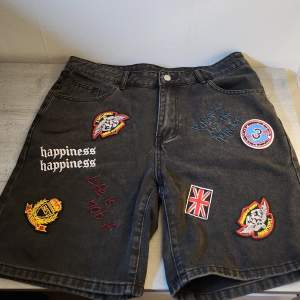 Säljer ett par svarta jeansshorts med coola patches och broderier. De har en normal passform och är perfekta för en avslappnad stil. Shortsen har flera färgglada patches, inklusive en brittisk flagga och text som 'happiness'. De har också klassiska femficksdetaljer och en knappgylf.