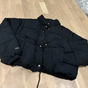 Svart pufferjacka - Säljer en svart pufferjacka som är perfekt för kalla dagar. Jackan har en quiltad design med långa ärmar och knappar framtill. Den är i bra skick och har en normal passform. Perfekt för både höst och vinter!