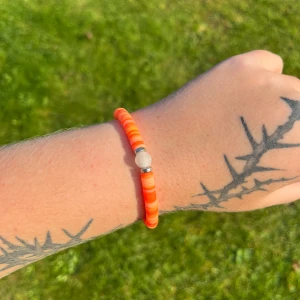 Orange armband med pärlor - Säljer ett snyggt orange armband med små, platta pärlor. Armbandet har en enkel och stilren design med en silverfärgad detalj i mitten. Perfekt för att liva upp vilken outfit som helst! Passar bra till sommarens alla äventyr. 🌞
