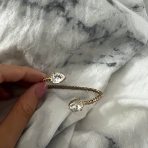 Guldarmband med kristaller - Säljer ett elegant guldarmband med en tvinnad design. Armbandet har två stora, gnistrande kristaller i ändarna som ger en lyxig känsla. Perfekt för att piffa upp vilken outfit som helst!INTRESSEKOLL