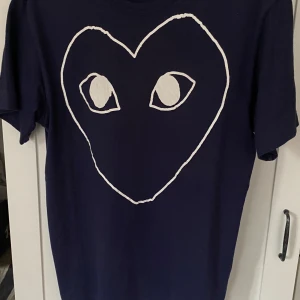 Mörkblå t-shirt med hjärtmotiv - Säljer en snygg mörkblå t-shirt från Comme des Garçons med ett coolt hjärtmotiv med ögon på framsidan. T-shirten är i storlek L och är gjord av 100% bomull. Perfekt för en avslappnad stil!