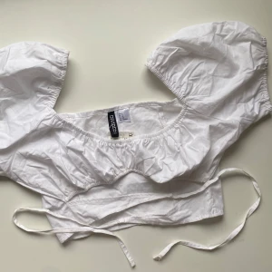 Vit kortärmad topp från Divided - Säljer en superfin vit kortärmad topp från Divided. Den har puffiga ärmar och en söt knytning framtill. Perfekt för sommardagar eller en festlig kväll. Toppen är helt ny med prislapp kvar.