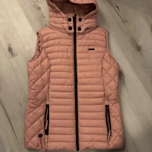 Rosa quiltad väst från Marikoo - Säljer en superfin rosa quiltad väst från Marikoo i storlek M. Västen har en praktisk dragkedja och en mysig huva. Perfekt för höst och vår när du vill hålla dig varm men ändå se snygg ut. Den är i mycket bra skick och redo för nya äventyr!