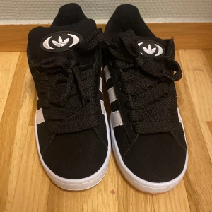 Svartac Adidas Campus sneakers - Säljer ett par snygga svarta Adidas Campus sneakers i mycket bra skick. De har den klassiska tre-randiga designen i vitt på sidan och en vit sula. Skorna är gjorda i mocka och har en bekväm passform med snörning. Perfekta för både vardag och fest!